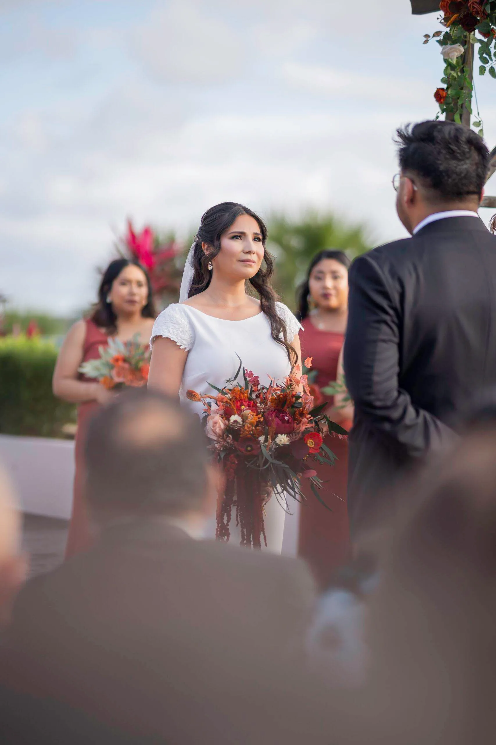 A bride holding a bouquet during an outdoor wedding ceremony, with guests softly blurred in the foreground.