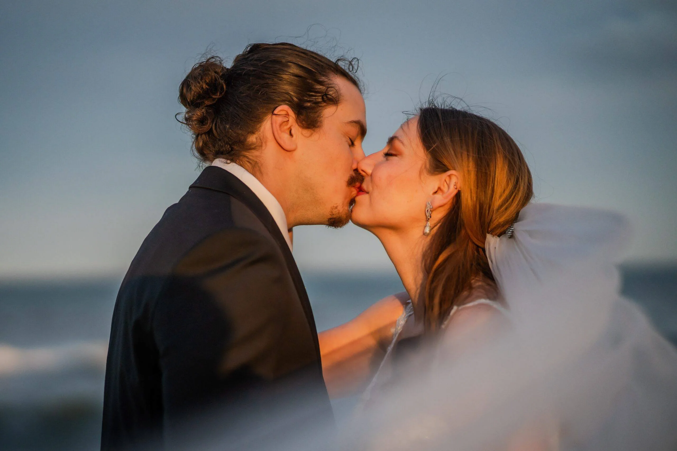 A bride and groom kissing with cool evening sky behind them.