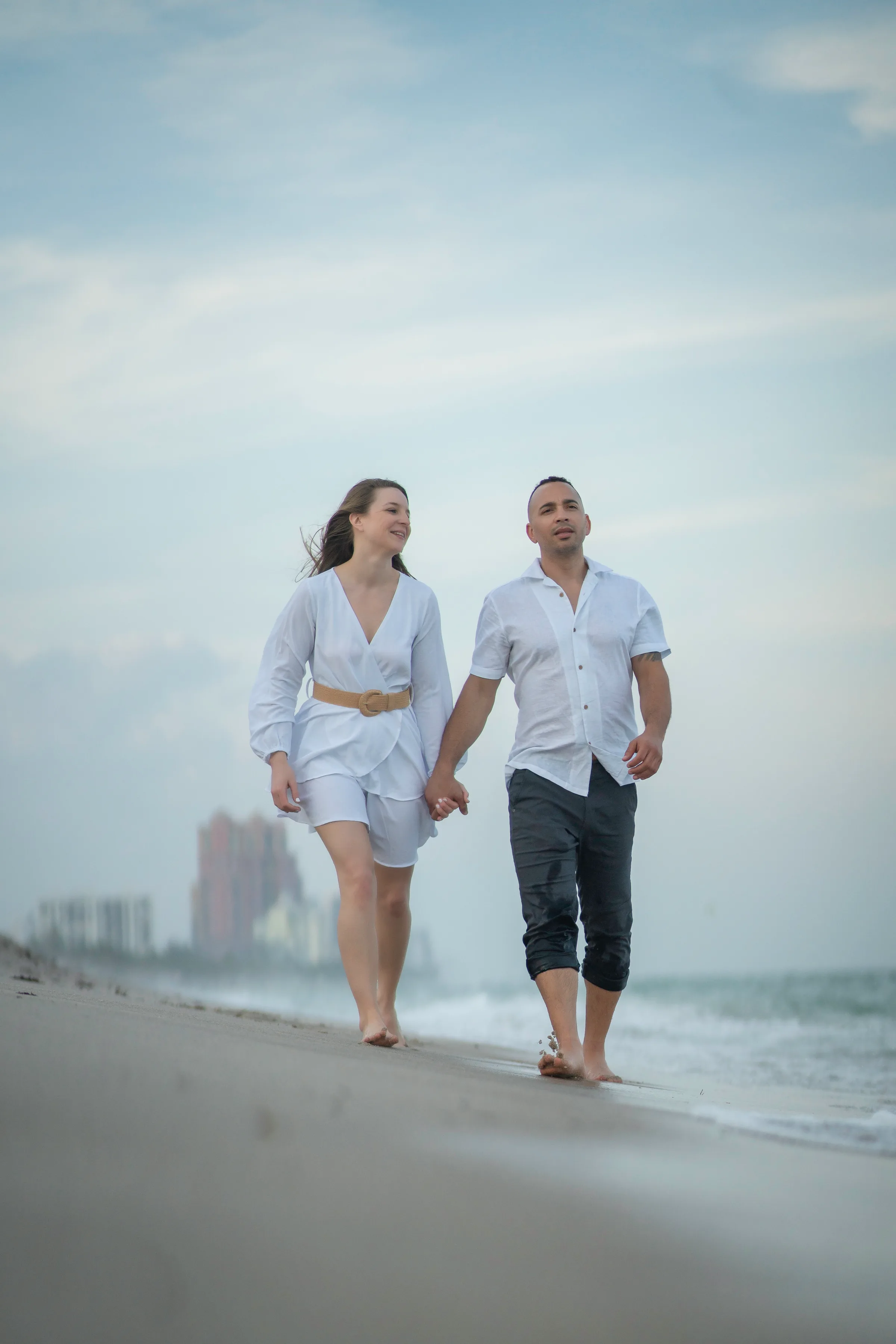 A couple walking barefoot along the shoreline in light-colored clothes.