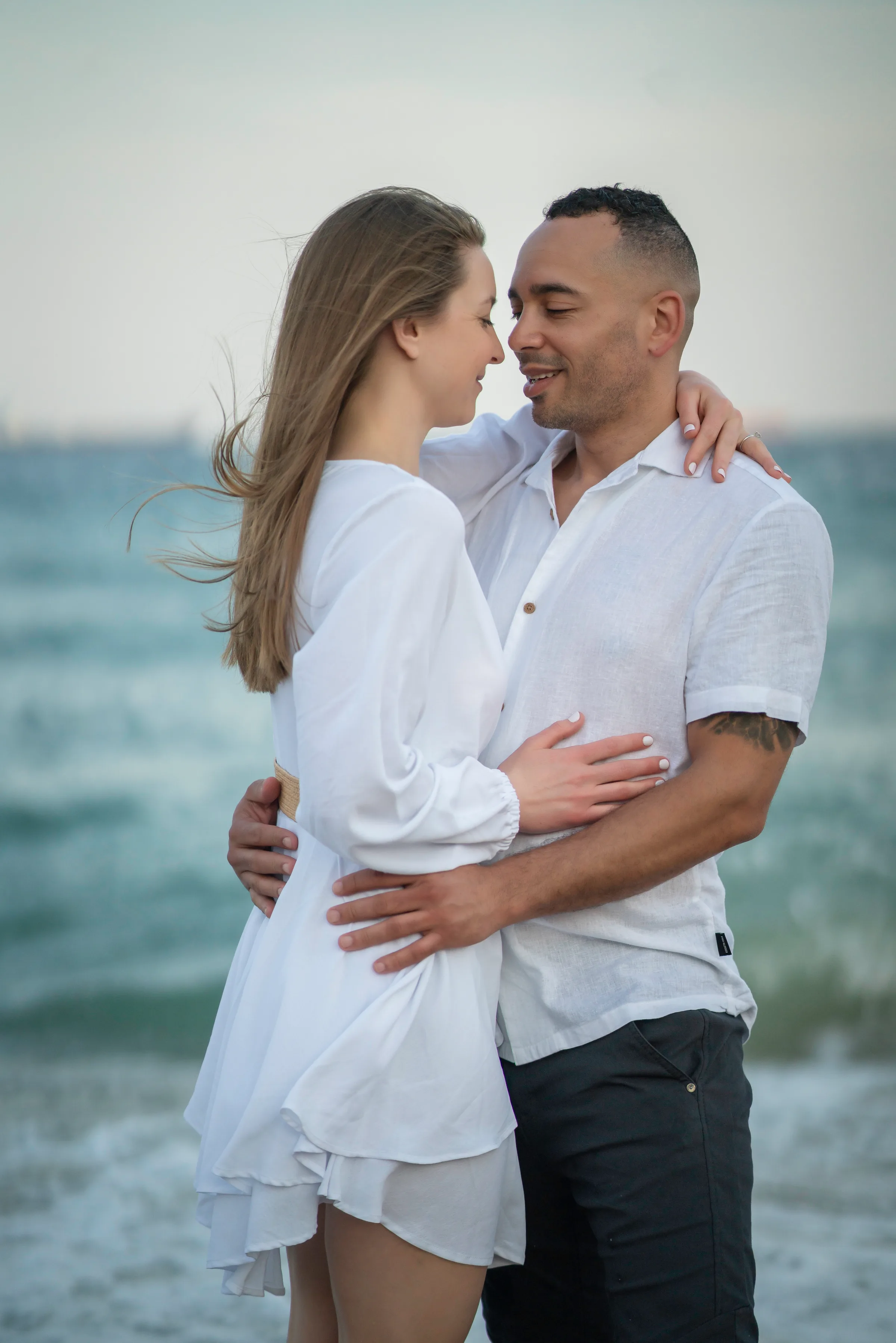A couple embracing beside the ocean, with one partner wearing a light, flowing dress.