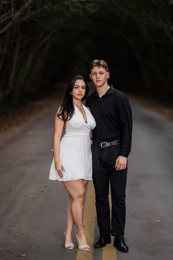 A man and a woman are standing together on the street. The man is wearing black pants while the woman is wearing white dress. They both have smiles on their face which indicates that they are happy.