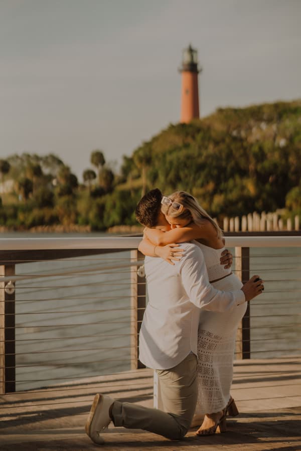 A couple embracing on a wooden pier with lighthouse in the background.