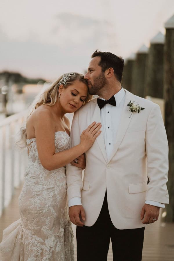 A man wearing a black bow tie stands next to a woman wearing a white dress with his hands on her shoulders.