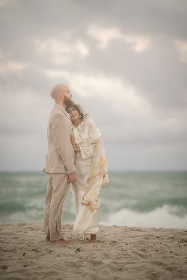 A man and a woman standing on the beach holding each other with their backs to the ocean.