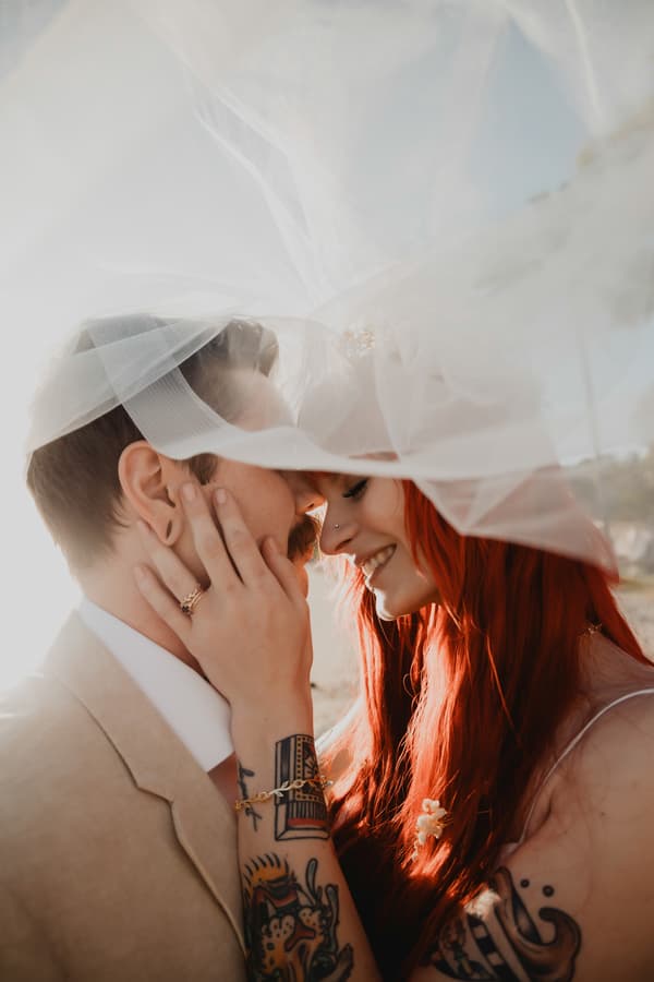 A man and a woman are kissing under a white veil. The man has tattoos on his arm and the woman has red hair. They are standing outside in what appears to be daytime.