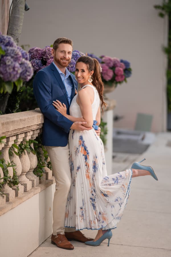 A man and woman are posing together with flowers behind them. The man is wearing a blue jacket and the woman is wearing a white dress with a blue shoe on her foot. They are standing in front of a wall that has purple flowers on it.