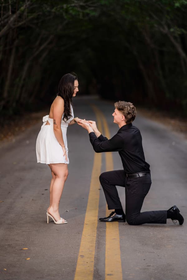 A man kneeling on the ground with a woman standing next to him. They are both wearing black clothing and have their hands together. The road they are on is dark and there are trees in the background.