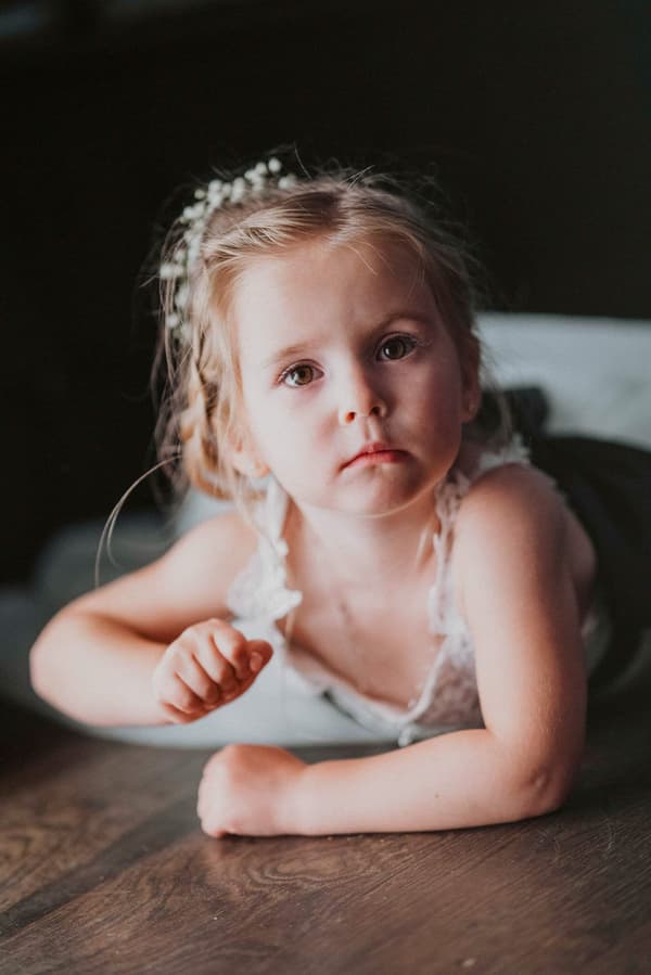A young girl with blonde hair is sitting on a wooden table looking at the camera.