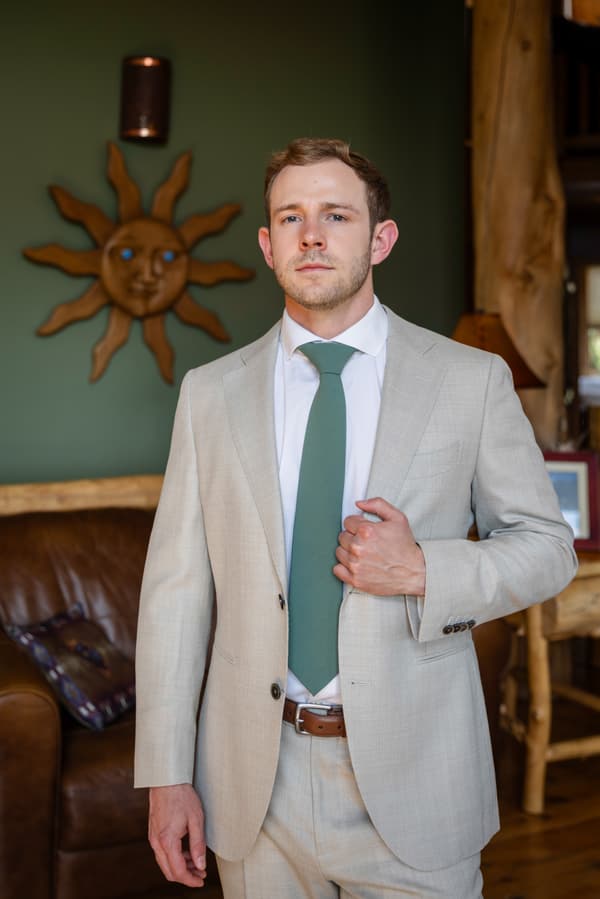 A man wearing a grey suit with a green tie stands in front of a sun shaped decoration on the wall.
