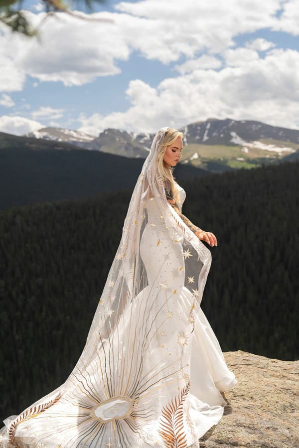 A woman wearing a white dress with gold accents stands on the edge of a cliff looking out over the mountains.