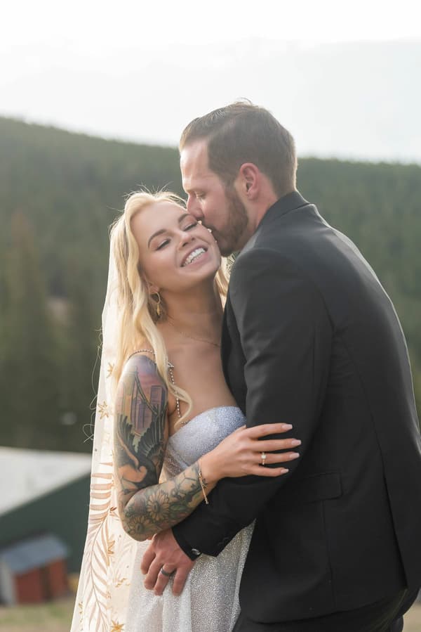 A man and a woman are standing together with the woman wearing a dress and tattoo on her arm. The man is holding the woman in his arms. They are both smiling at each other.