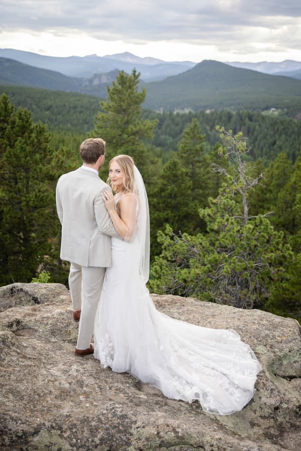 A bride and groom stand on a rock outcropping overlooking the mountainside.