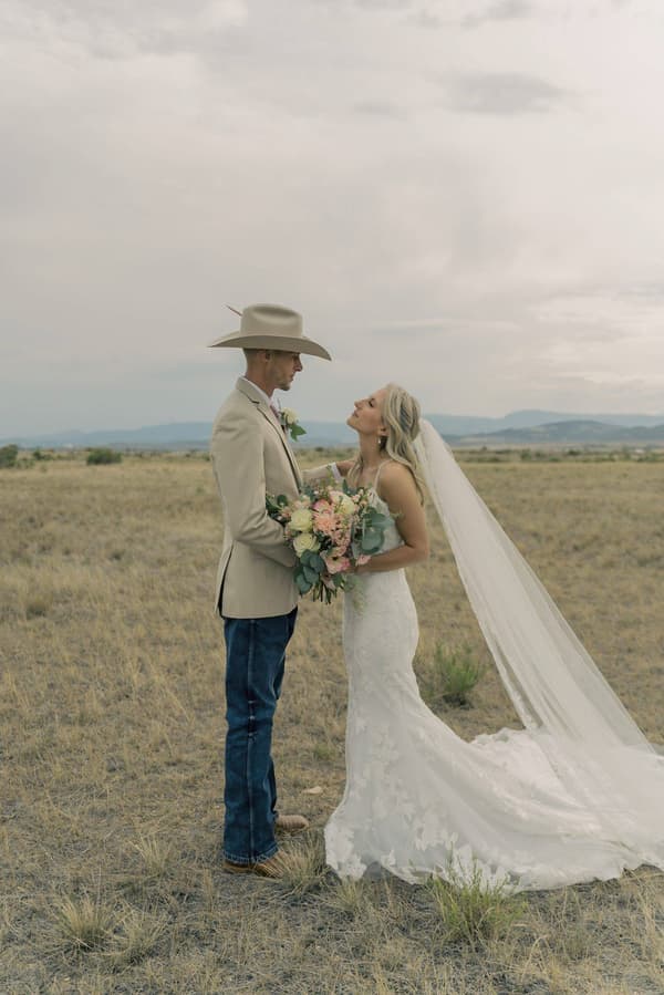 * A man and a woman are standing together on the grass. The man is wearing a tan hat and holding flowers. The woman is wearing a white dress with a veil over her head. They appear to be newly married.