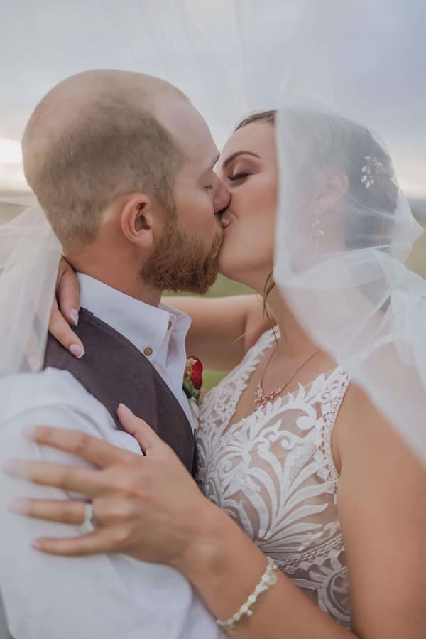 A man and a woman are kissing under a white veil. The man is wearing a vest with a red rose on it. They are standing outside in the sunshine.