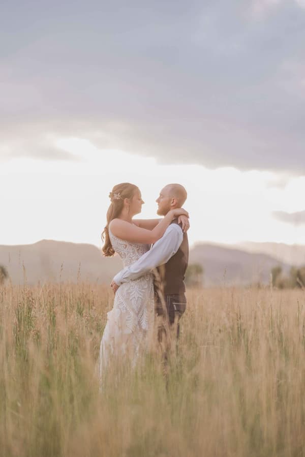 A man and woman are standing together in a field of tall grass with mountains in the background. The sky is cloudy and the sun is shining through.