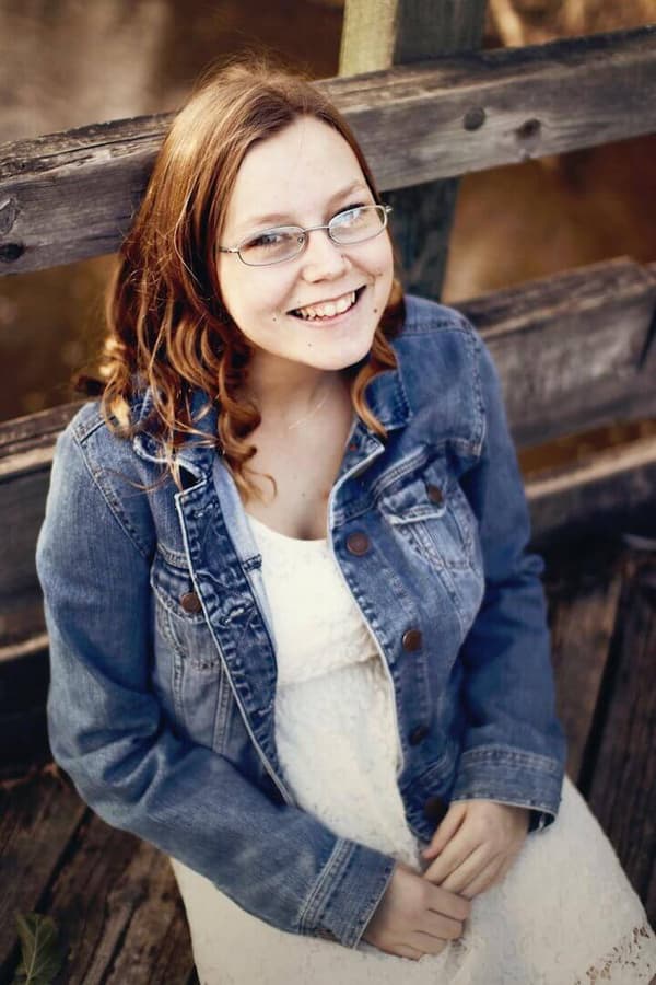 A woman wearing glasses is sitting on a wooden bench with her hands folded across her chest. She has red hair and is smiling at the camera. The background of the image is blurred but appears to be an outdoor setting with trees and bushes. The mood of the photo is cheerful and relaxed, as if the woman is enjoying a quiet
