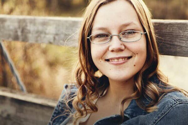 A girl wearing glasses is sitting on a wooden bench with her mouth closed.
