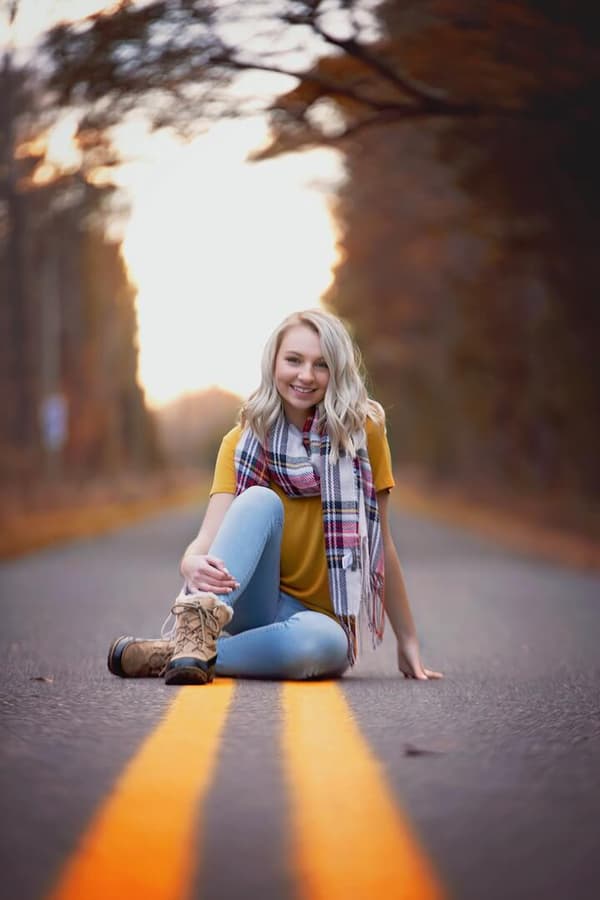 A woman is sitting on the road with a yellow shirt and blue jeans. She has brown boots on her feet. The sun is going down behind her.