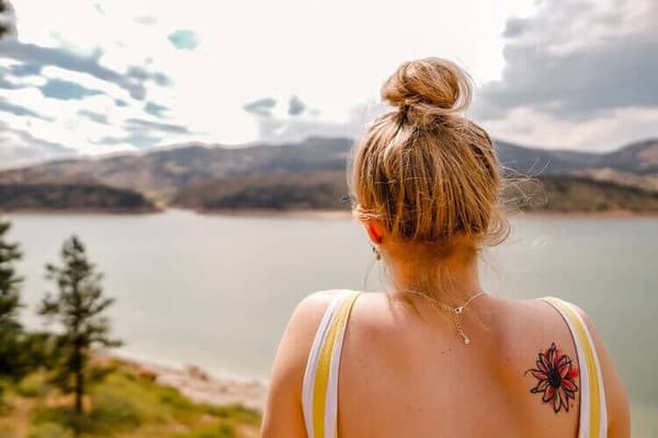 A woman with a red flower tattoo on her back is looking out over the water at a mountain range.