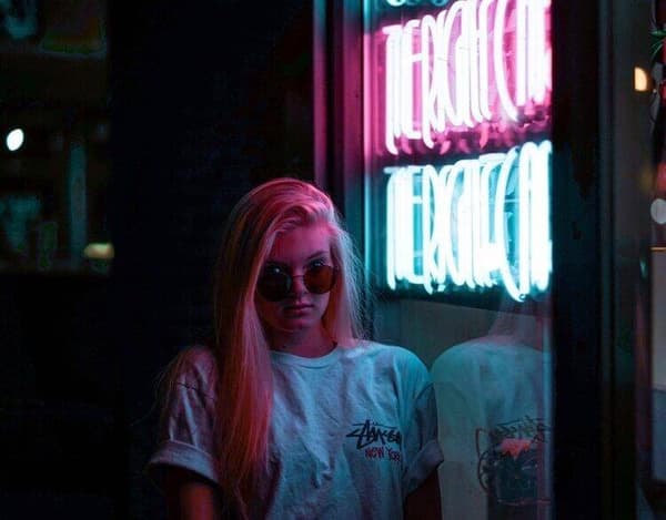A girl with sunglasses on her face is standing outside a bar that says "Vanity". The background of the photo is dark.